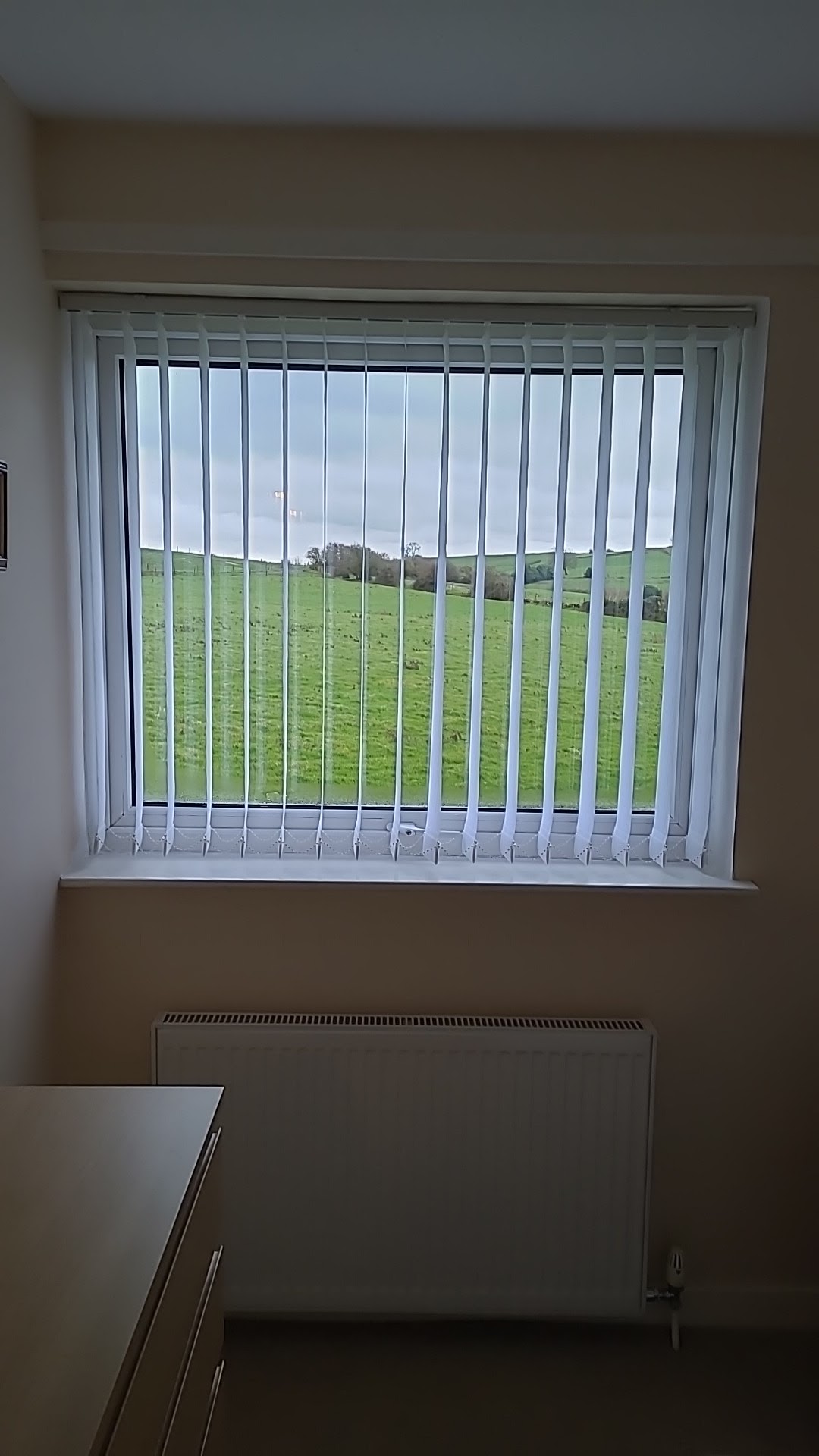 White vertical blinds fitted across a window overlooking green fields